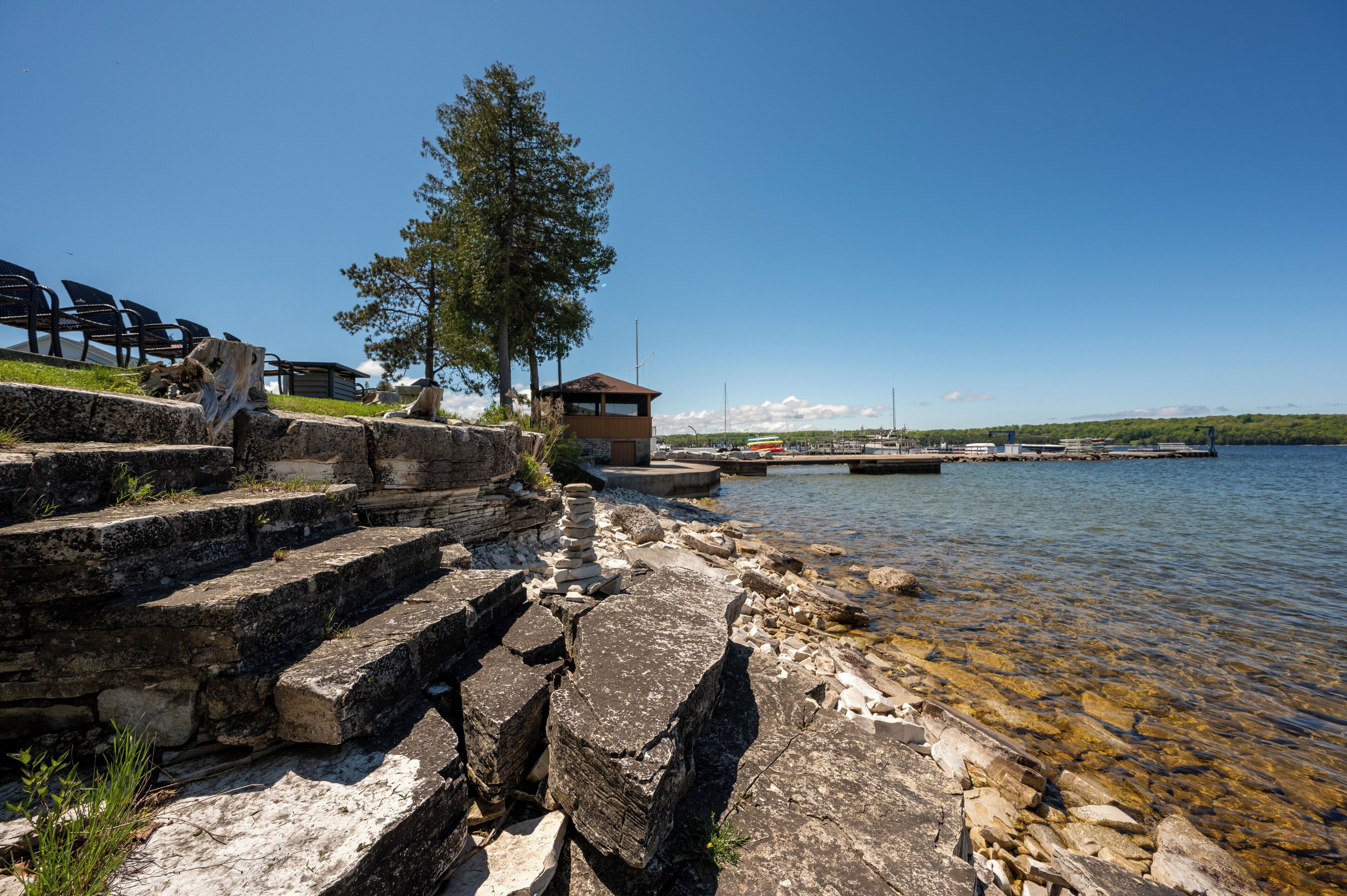 Una escalera de piedra conduce a una cabaña de madera junto a un lago, con agua cristalina y un muelle al fondo.