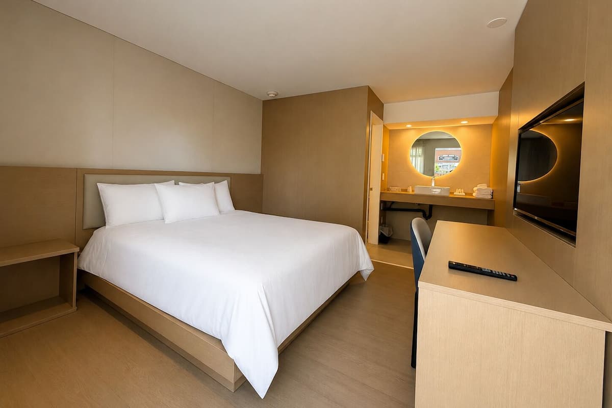 A hotel room with a bed, a desk, a chair, a TV, and a bathroom with a mirror and sink.