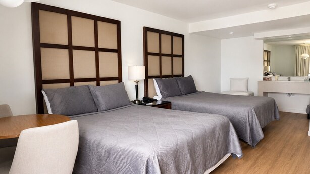 Habitación de hotel con dos camas, un escritorio, una silla, una mesita de noche, una lámpara, un espejo y piso de madera.