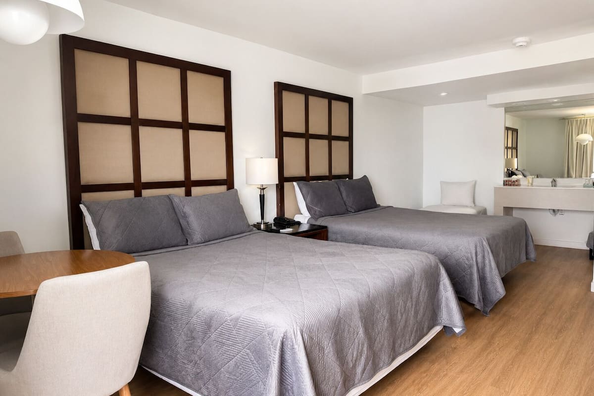 A hotel room with two beds, a desk, a chair, a nightstand, a lamp, a mirror, and a wooden floor.