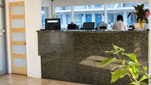 Un mostrador de recepción con una computadora, un teléfono y una impresora. Se ve una ventana con vista a un edificio.