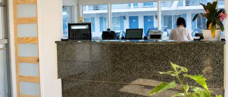 Un mostrador de recepción con una computadora, un teléfono y una impresora. Se ve una ventana con vista a un edificio.
