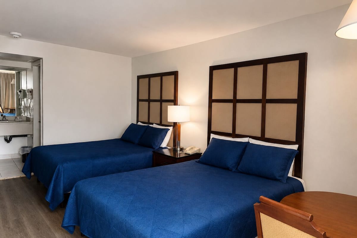 A hotel room with two beds, a bedside table with a lamp, a chair, and a mirror.