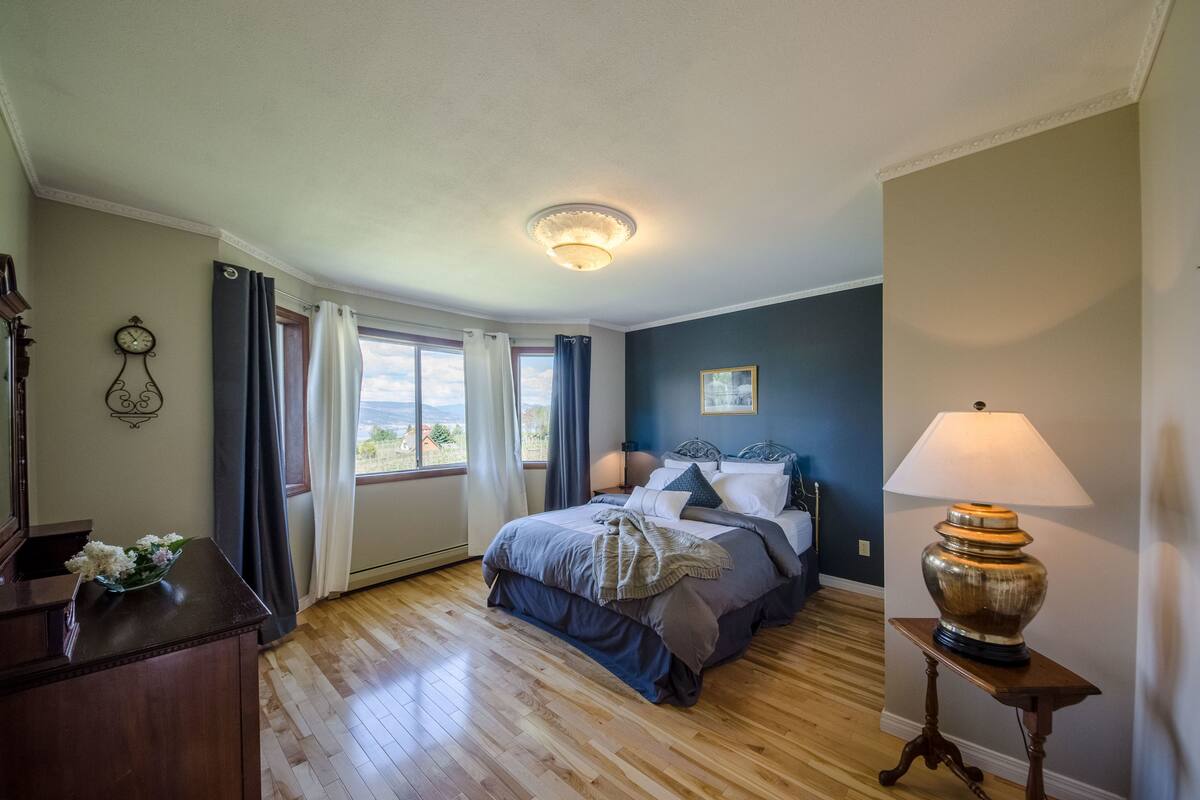 A bedroom with a bed, a bedside table, a lamp, a clock, and a window with curtains.