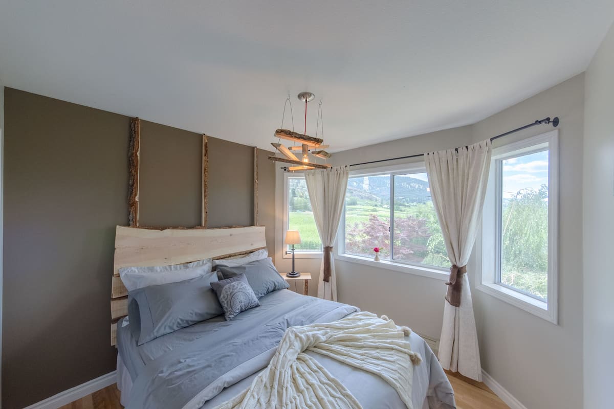 A bedroom with a wooden headboard, a bed with grey bedding, a white throw blanket, a bedside table with a lamp, and a window with curtains.