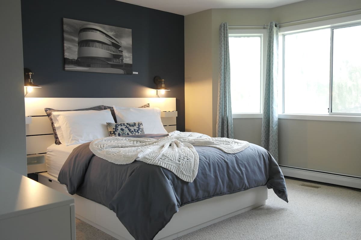 A modern bedroom with a bed, bedside tables, a nightstand, a wall-mounted lamp, and a window with curtains.