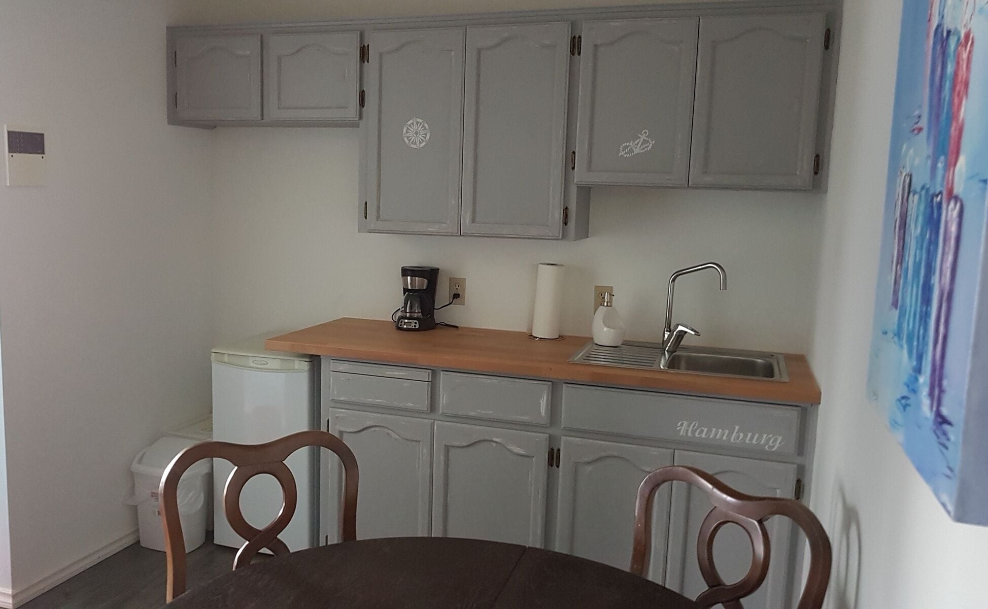 A compact kitchen with grey cabinets, a wooden countertop, a coffee maker, a sink, and a chair.