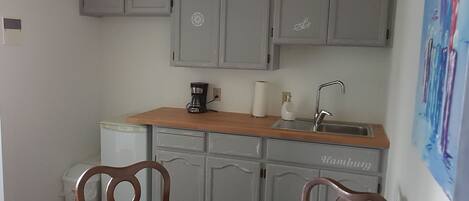A compact kitchen with grey cabinets, a wooden countertop, a coffee maker, a sink, and a chair.