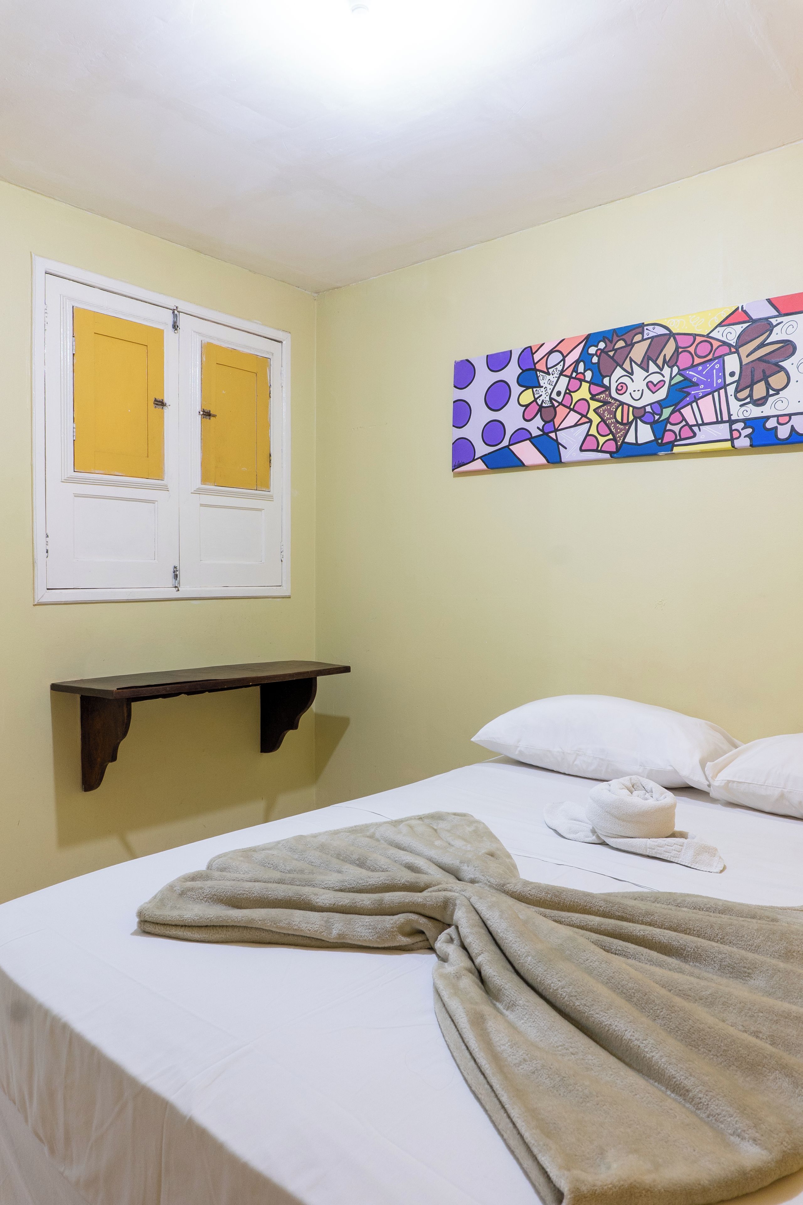 A room with a bed, a window with yellow curtains, a wall-mounted shelf, and a colorful abstract painting.