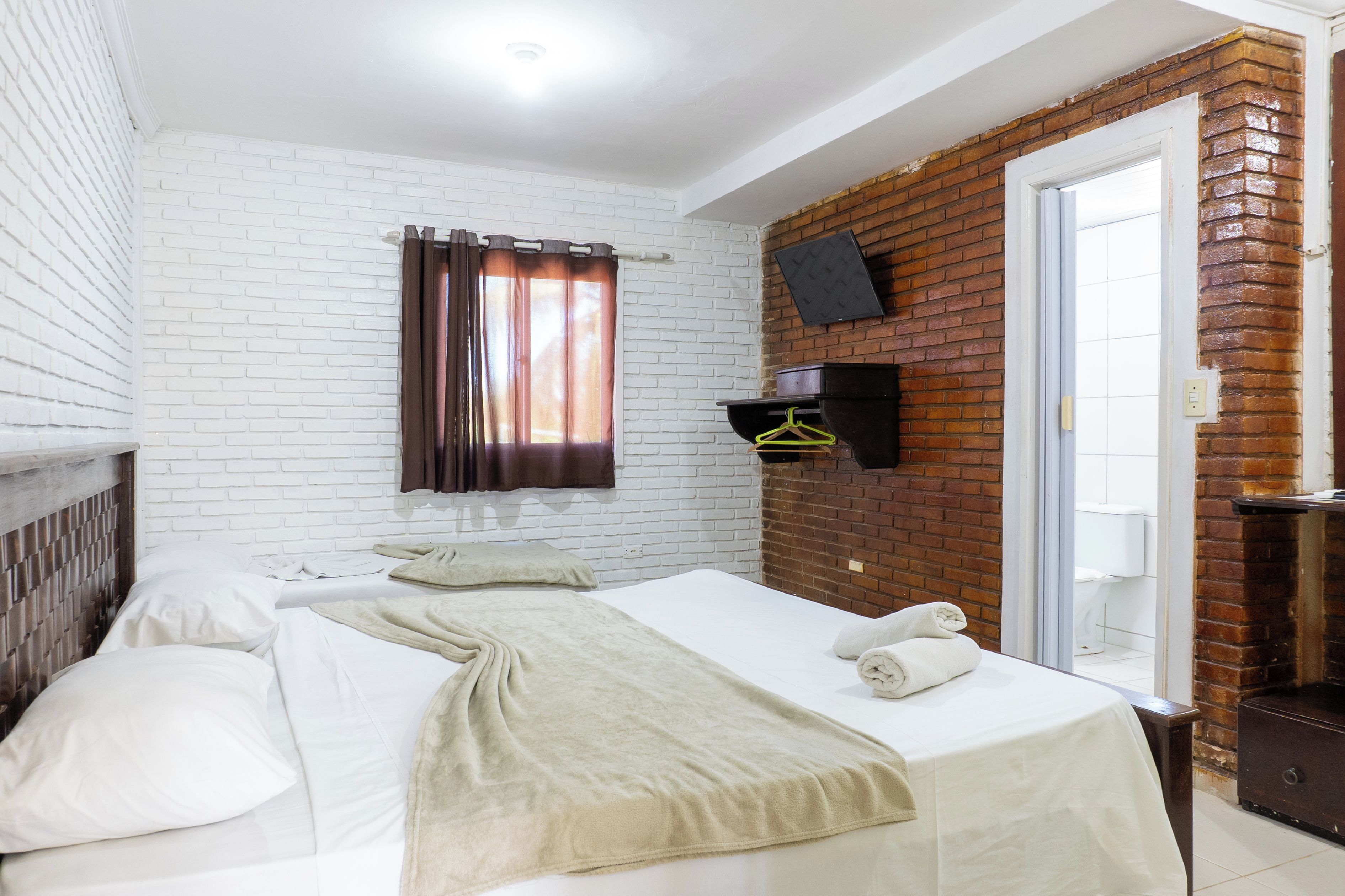 Una habitación de hotel con pared de ladrillo blanco, cama con ropa blanca, cortina marrón, estante pequeño negro y puerta que da a un baño.