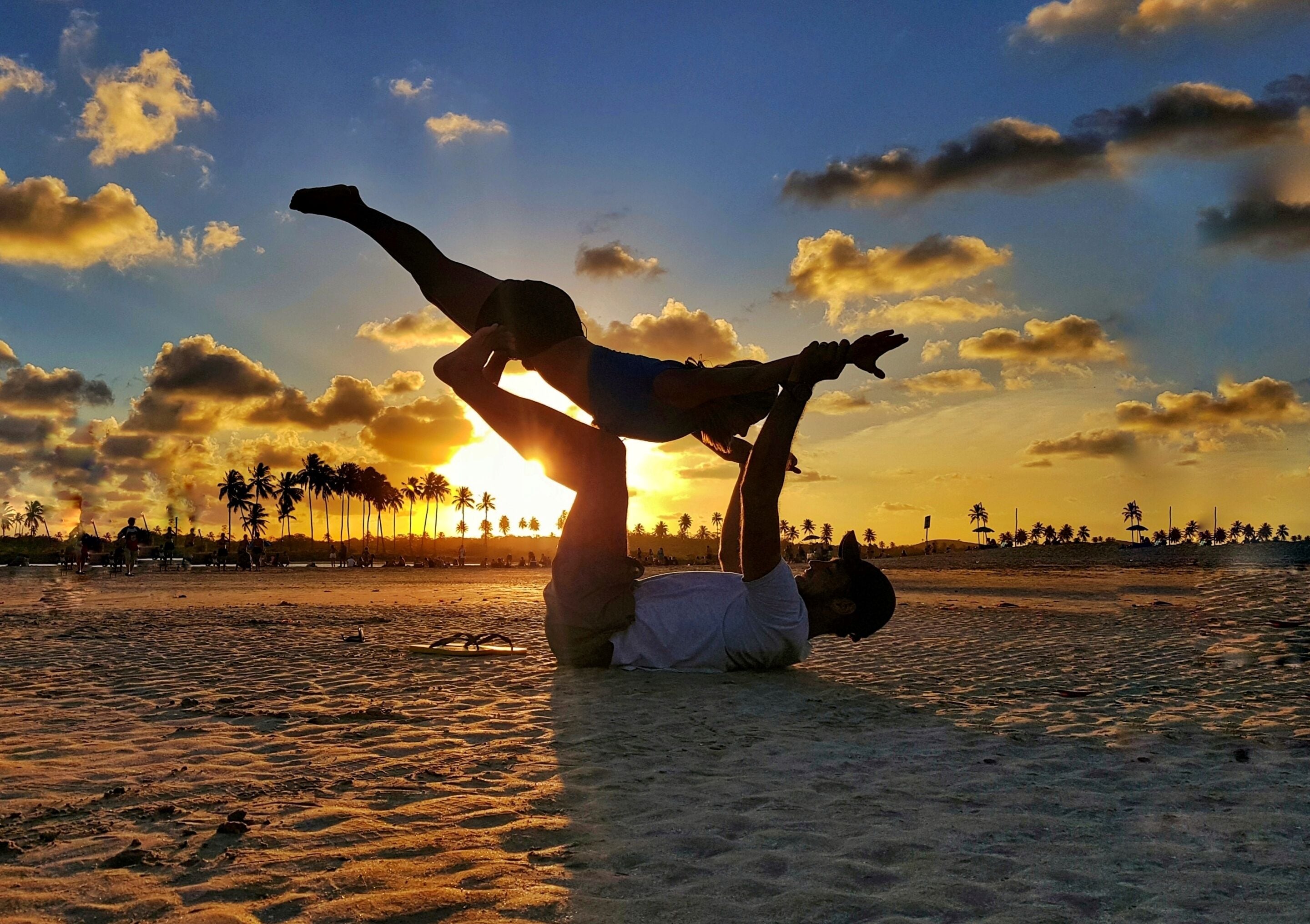 A couple performing an acrobatic stunt on a beach with palm trees and a sunset in the background.