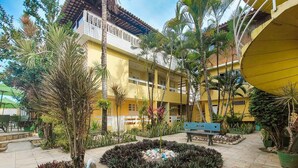 A courtyard with a yellow building, palm trees, and a bench. - Pousada Jardins do Porto (Ipojuca)