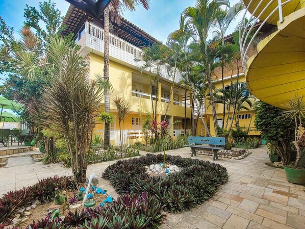 A courtyard with a yellow building, palm trees, and a bench. - Pousada Jardins do Porto (Ipojuca)