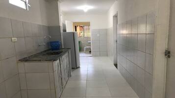 Un pasillo estrecho con un área de cocina a la izquierda, que incluye un lavamanos, una cesta azul y un refrigerador. El pasillo tiene paredes y suelo de baldosas blancas, que conducen a una puerta a la derecha.