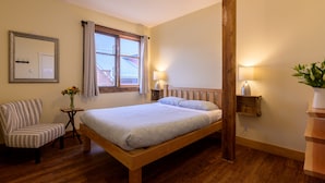 Une chambre à coucher avec un lit en bois, une chaise à rayures, une petite table sur laquelle est posé un vase de fleurs, et une fenêtre avec des rideaux.