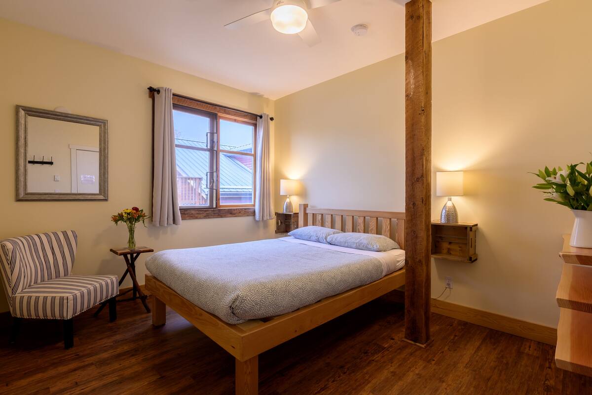 A bedroom with a wooden bed, a striped chair, a small table with a vase of flowers, and a window with curtains.