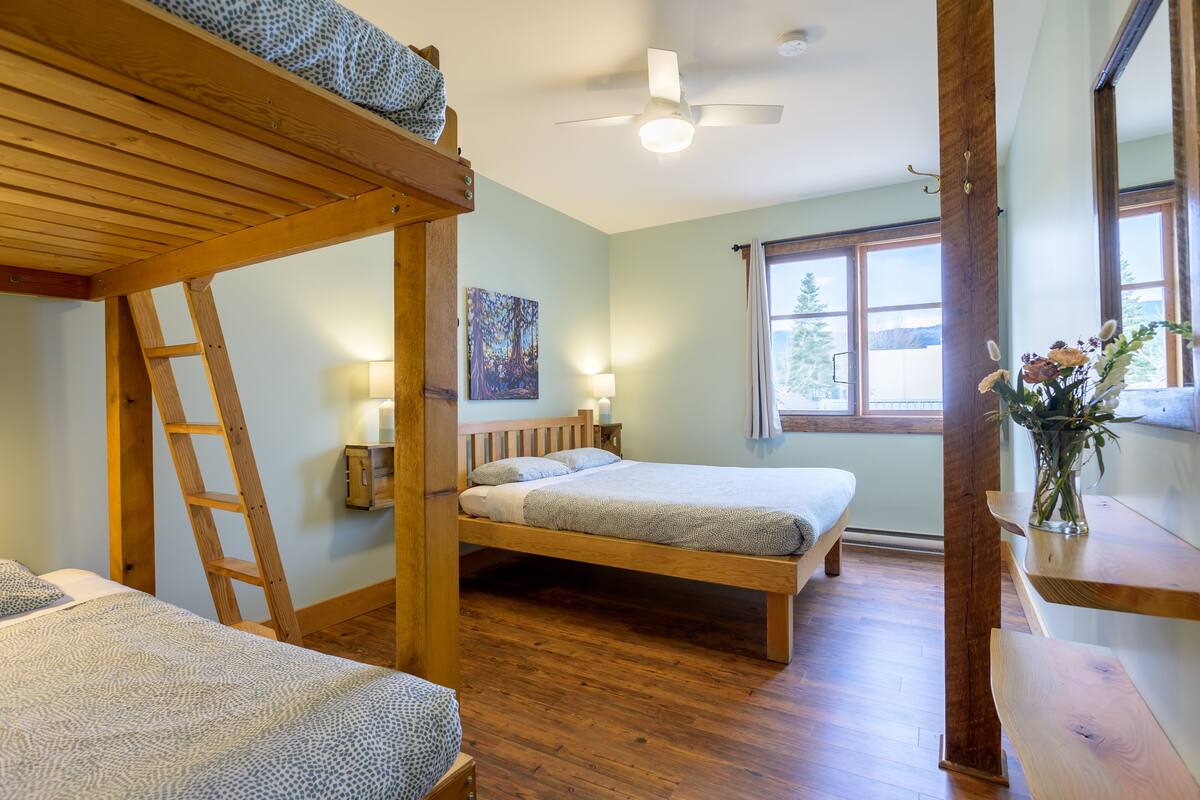 A room with two bunk beds, a wooden ladder, a ceiling fan, a window with a view of trees, and a vase with flowers on a shelf.