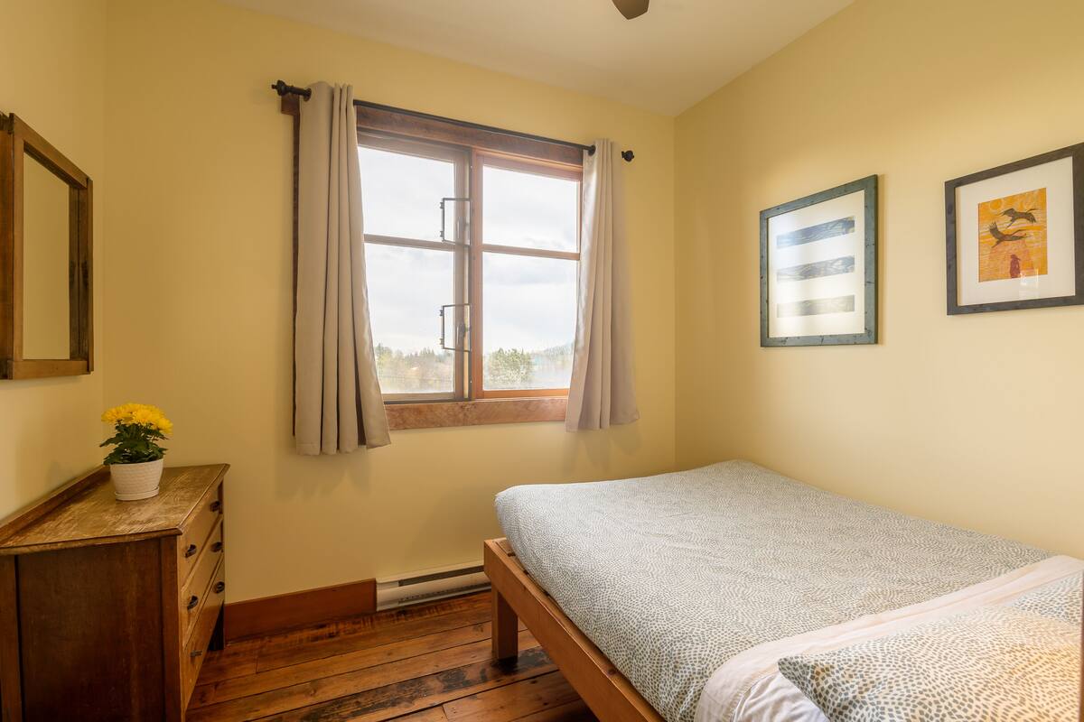 A bedroom with a wooden bed, a window with curtains, a dresser with a potted plant, and two framed pictures on the wall.