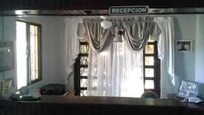 A reception area with a sign that reads "RECEPCION" above a counter. There is a window with curtains, a desk with a computer, and a chair. - Green House Aparta Hotel (Santo Domingo)