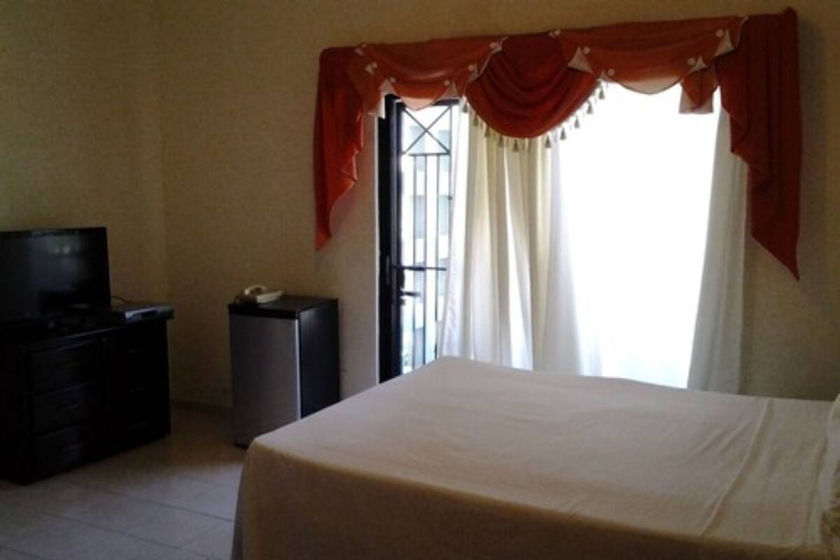 Une chambre à coucher avec un lit, une commode, un téléviseur et une fenêtre avec des rideaux.