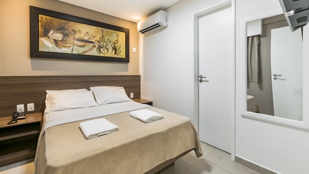 A hotel room with a bed, a painting on the wall, an air conditioning unit, a bedside table with a phone, and a door with a mirror. - VilaHotell Recife (Recife)