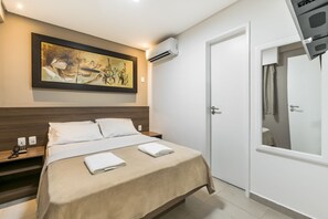 Habitación de hotel con cama, un cuadro en la pared, equipo de aire acondicionado, mesita de noche con teléfono y puerta con espejo.