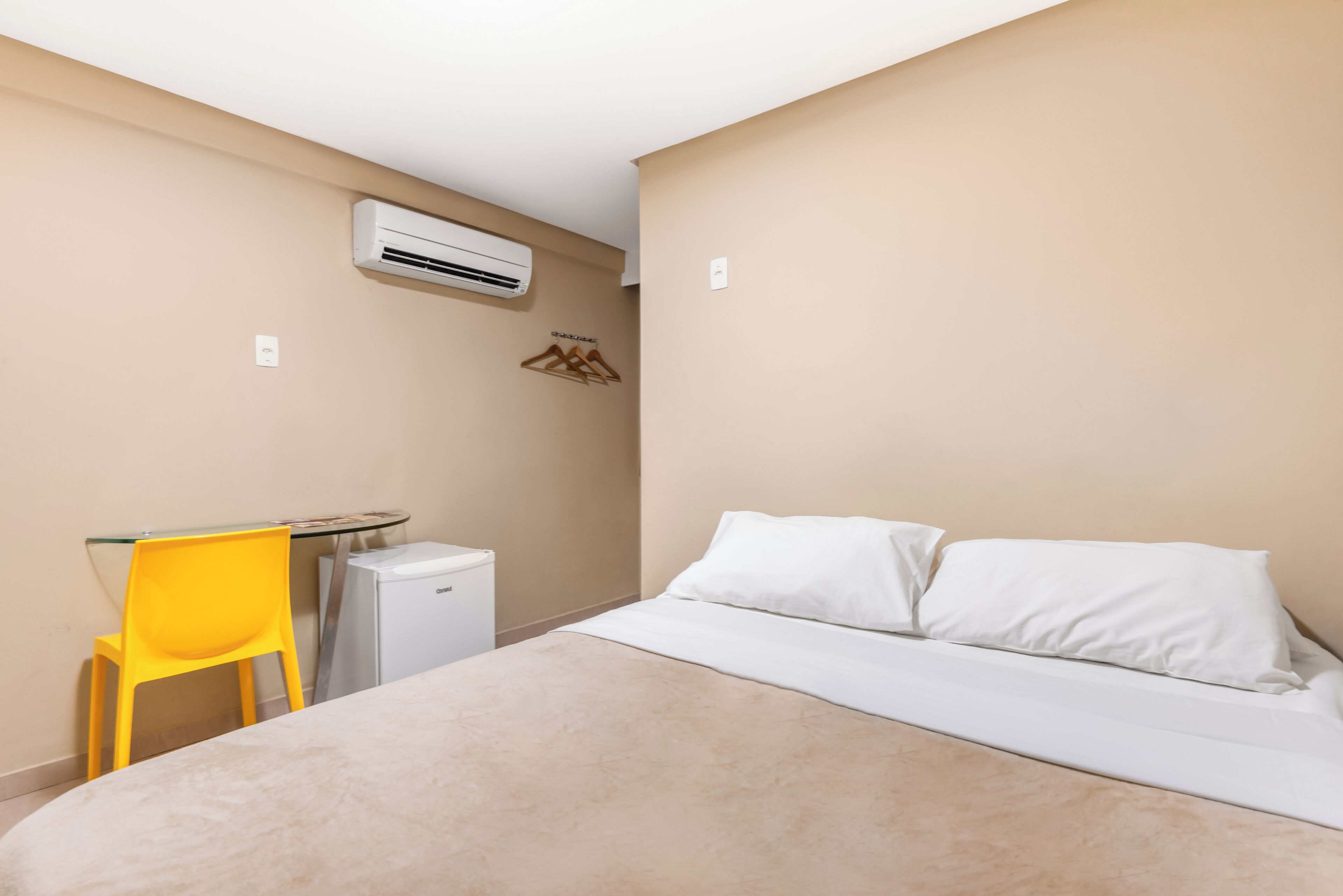 Hotellihuoneessa on sänky, pieni pöytä, keltainen tuoli, valkoinen ilmastointilaite ja seinään kiinnitetty lämmitin.