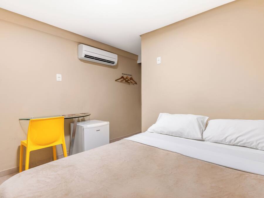 Habitación de hotel con cama, una mesita, una silla amarilla, un aire acondicionado blanco y un calentador fijado a la pared.