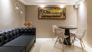 A modern living room with a black leather sofa, a round dining table with white chairs, and a large painting of a historical scene on the wall. - VilaHotell Recife (Recife)