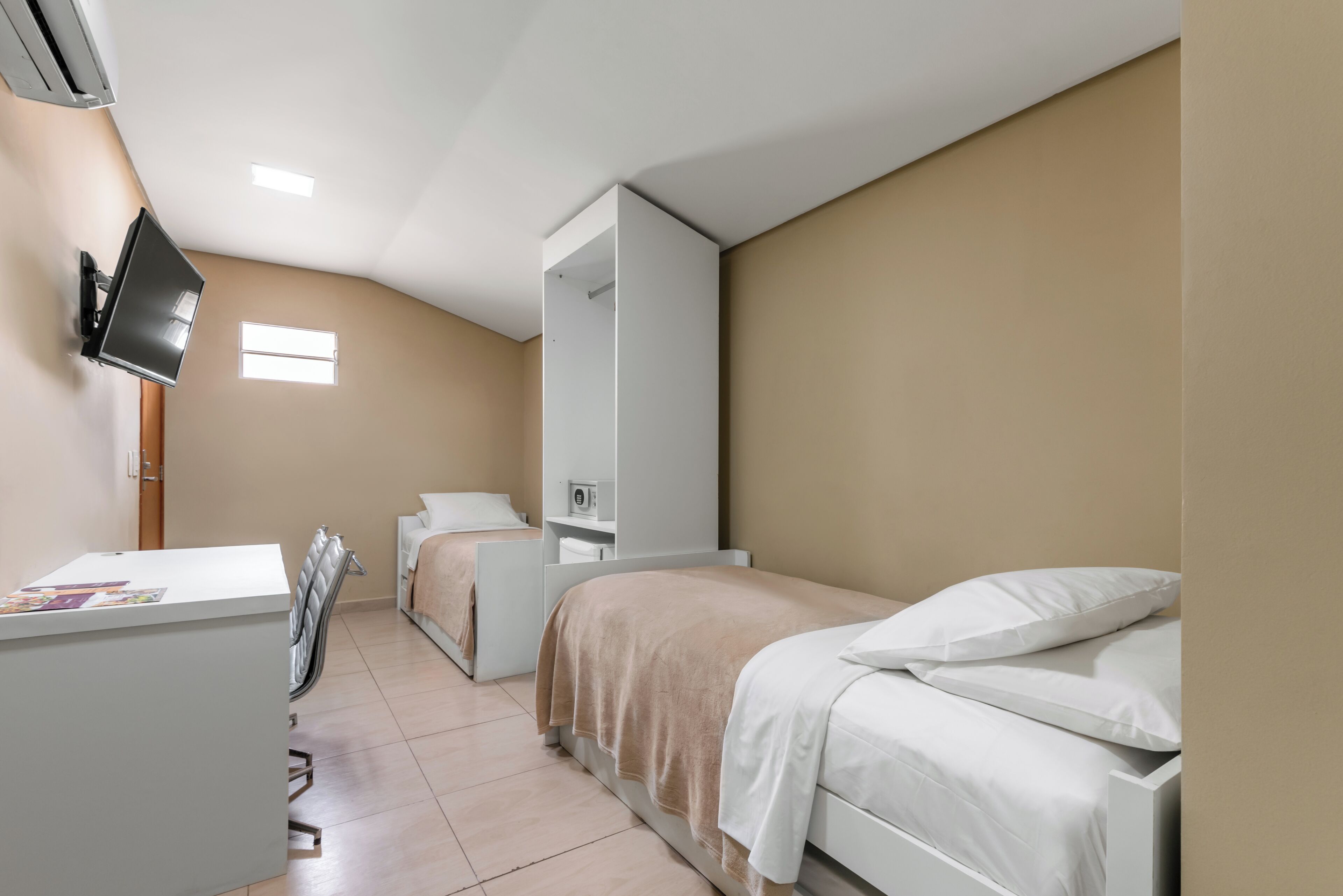 A hotel room with a desk, two beds, a TV, and a small window.
