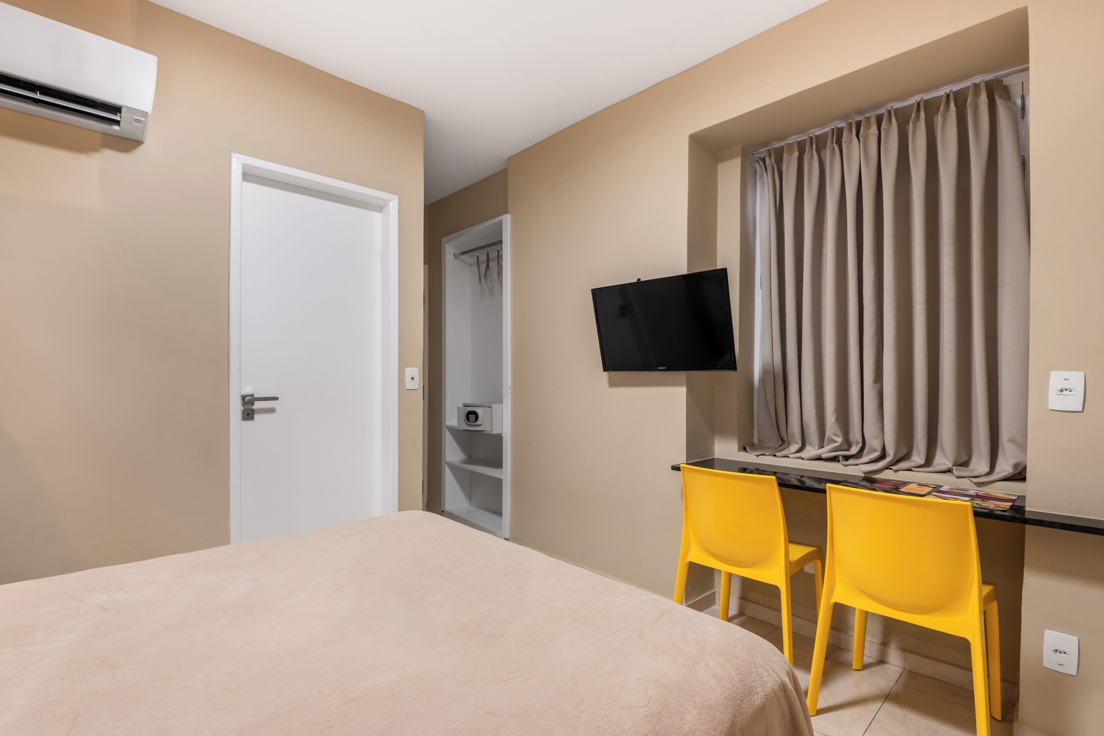 A bedroom with a bed, a desk with two chairs, a television, and an air conditioning unit.