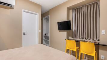A bedroom with a bed, a desk with two chairs, a television, and an air conditioning unit.