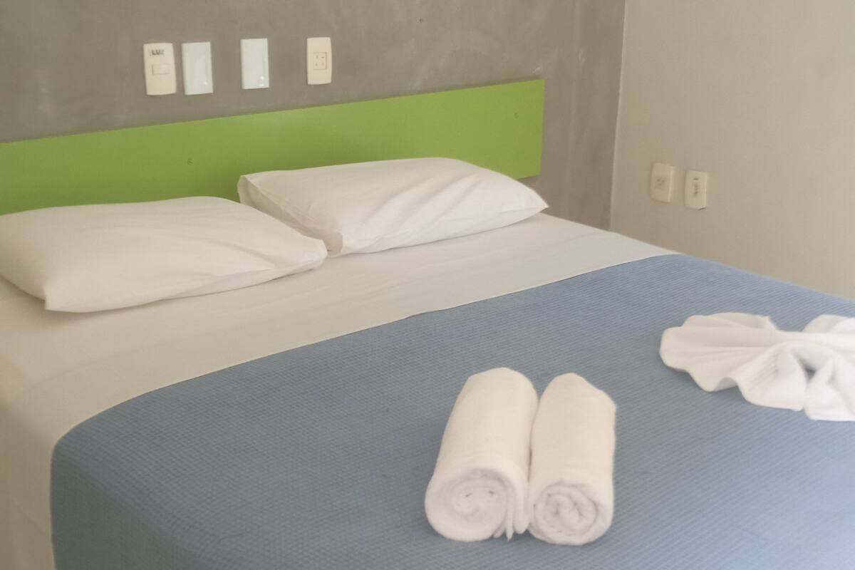 A bed with white pillows and a blue bedspread, two rolled towels on the bed, and a green headboard.