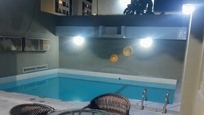 An outdoor pool area with a glass table and wicker chairs, surrounded by a low wall and a building with illuminated windows. - Pousada Boi Bumbá Praia (João Pessoa)