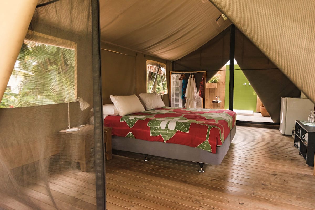 A bedroom with a bed, wooden flooring, a window with a view of greenery, and a door leading to another room.