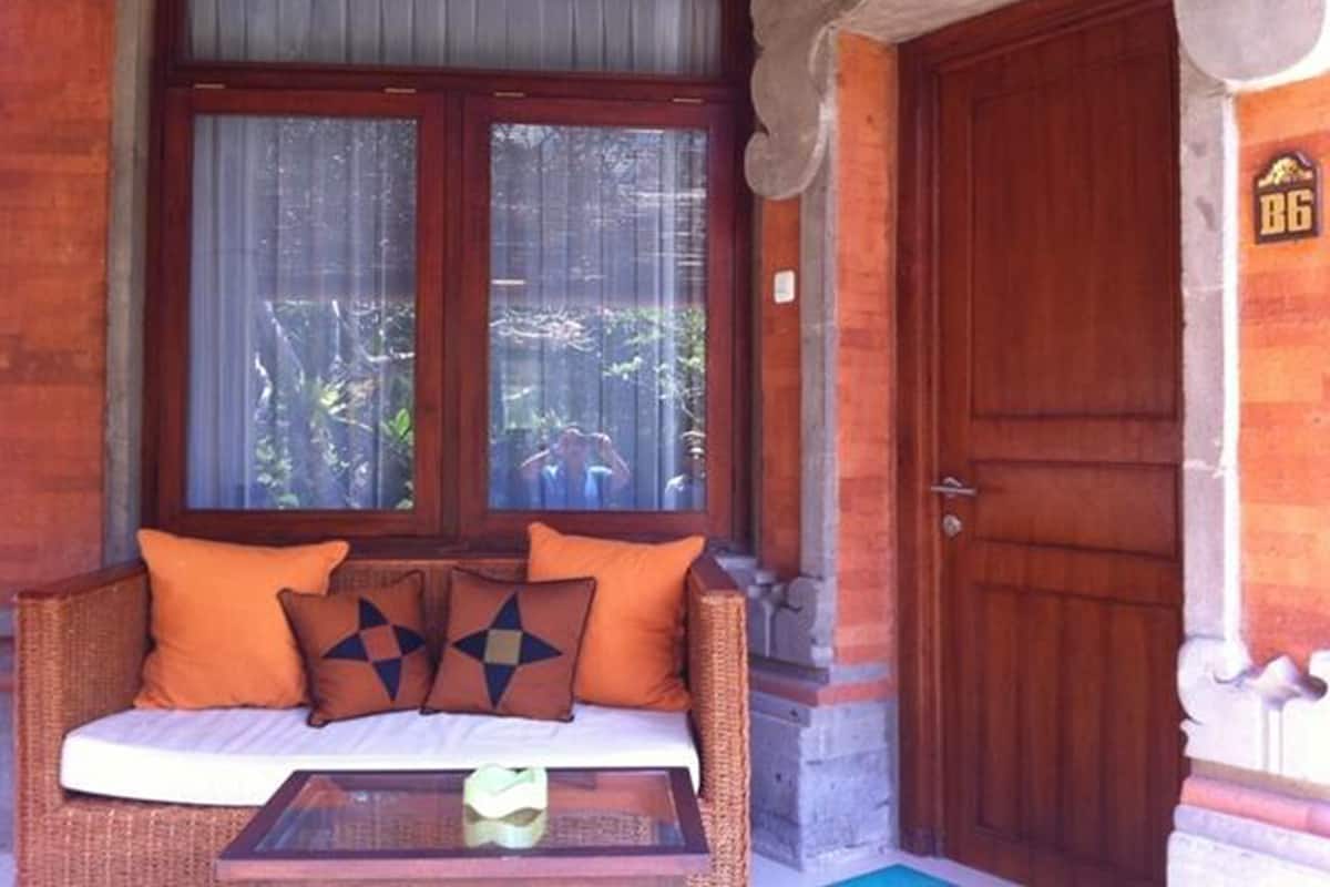 Um quarto com porta de madeira, janela com cortinas, sofá de vime com almofadas laranja e uma pequena mesa.