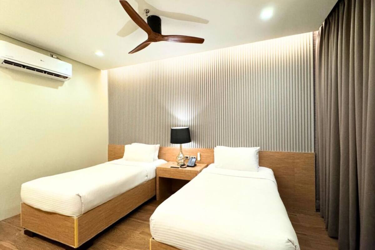 A hotel room with two single beds, a ceiling fan, an air conditioning unit, a bedside table with a lamp, and a curtain on the window.