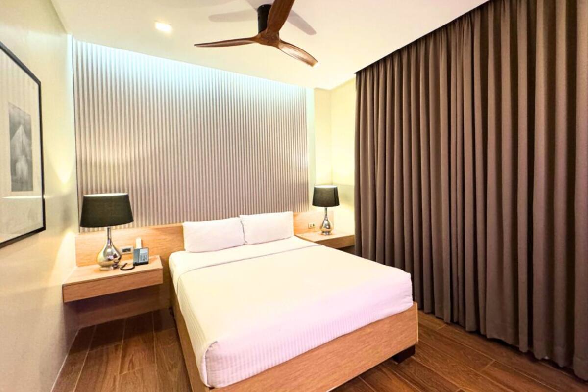 A hotel room with a large bed, two bedside tables with lamps, a ceiling fan, and a view of the outside through curtains.