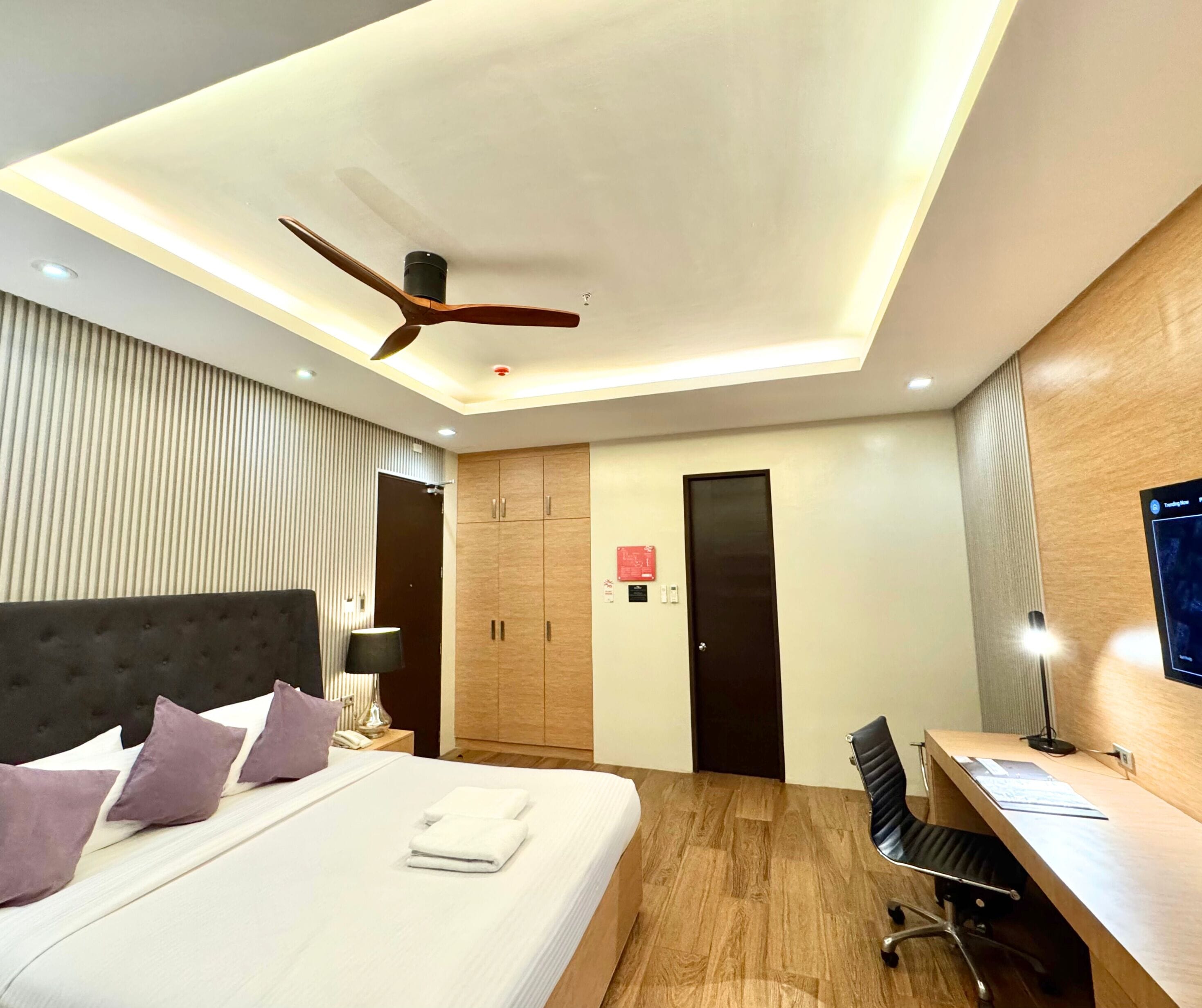A modern hotel room with a bed, desk, chair, wardrobe, and ceiling fan.