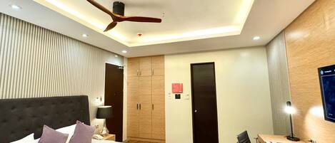 A modern hotel room with a bed, desk, chair, wardrobe, and ceiling fan.