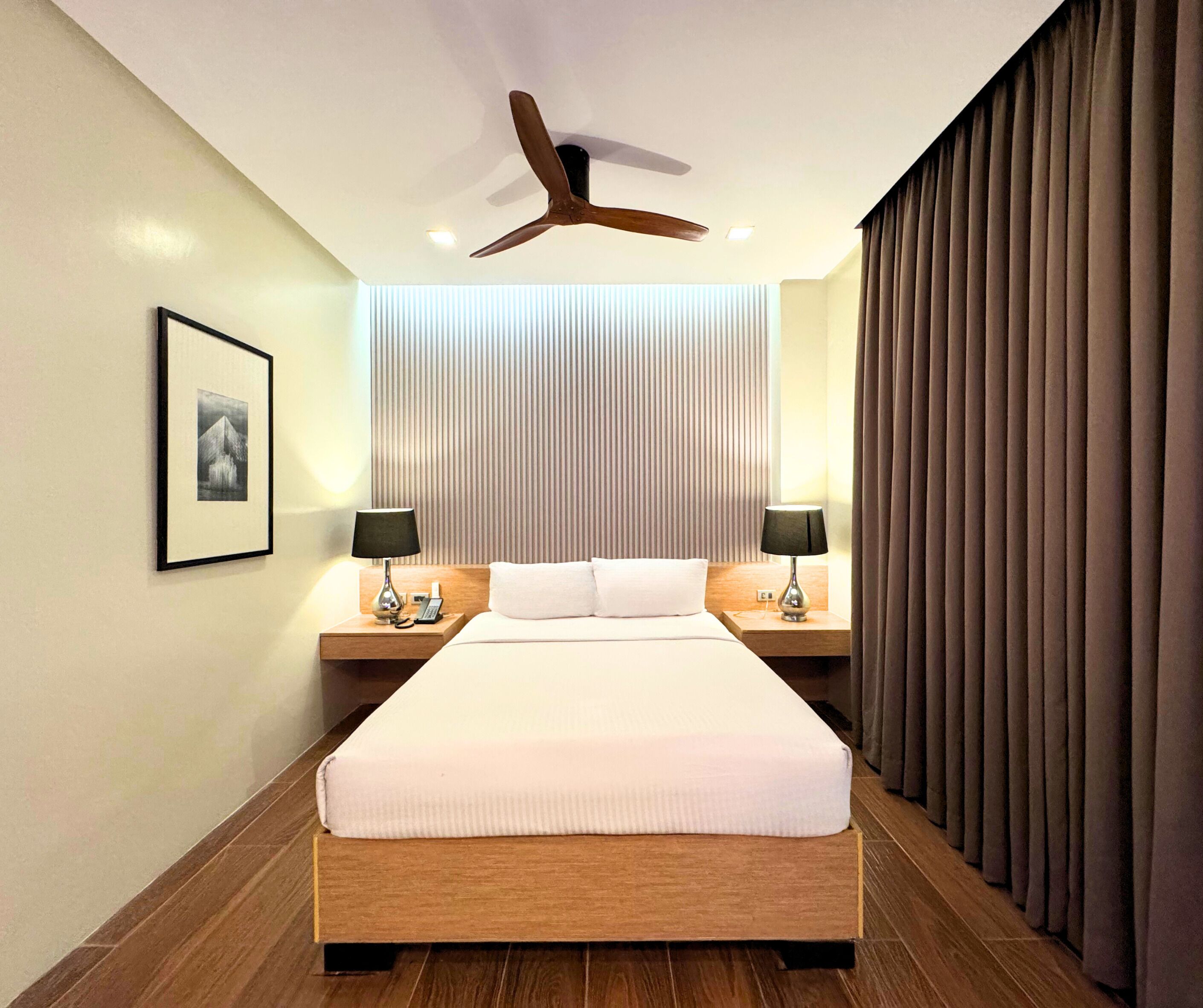Une chambre d’hôtel avec un grand lit, deux tables de chevet avec des lampes, un ventilateur de plafond, un tableau encadré au mur et une fenêtre avec des stores verticaux.