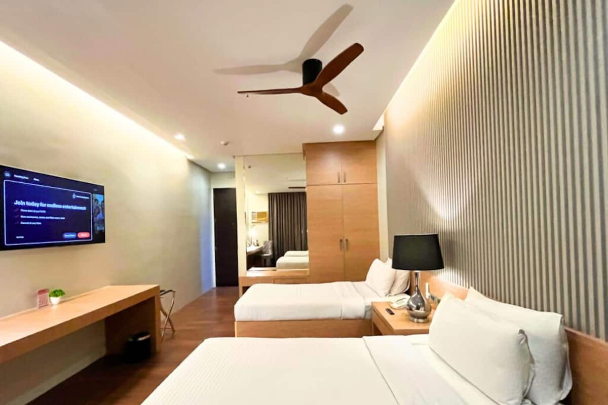 A hotel room with two beds, a desk, a TV, a ceiling fan, and a wall-mounted TV.