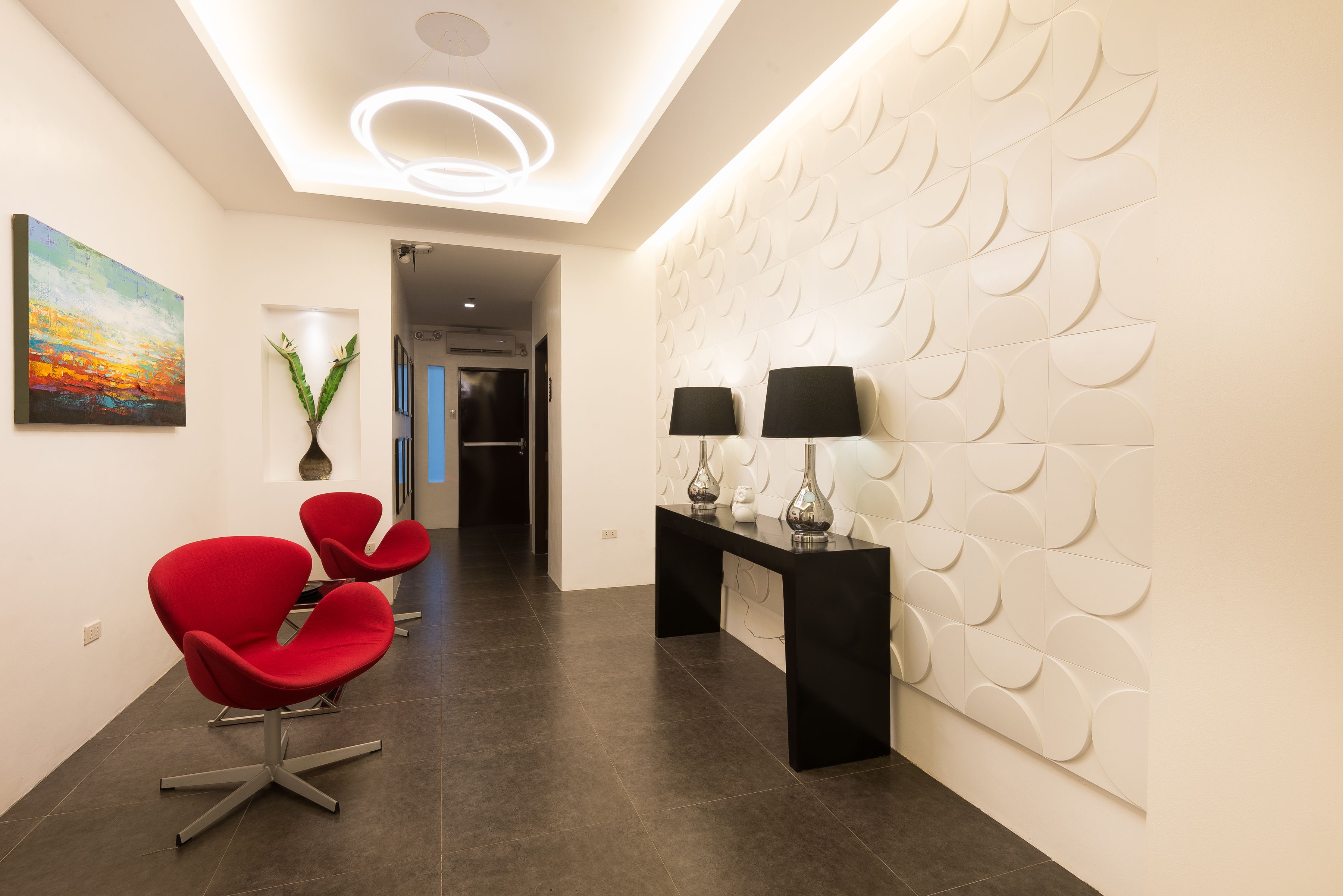 Un interior moderno con un mostrador de recepción, dos sillas rojas, una pared con un diseño de hojas y un cuadro colgado.