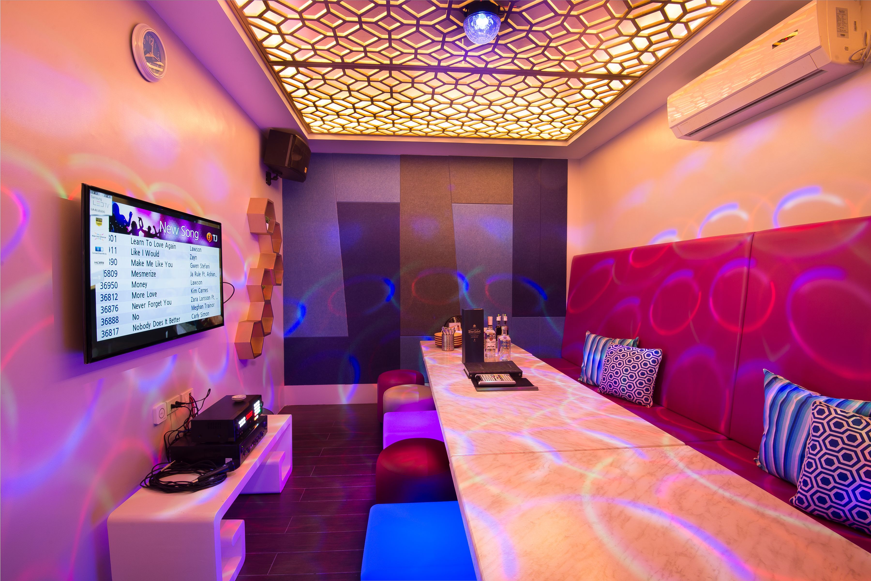 A modern lounge area with a digital screen displaying music, a ceiling with geometric lighting, and a sofa with patterned cushions.