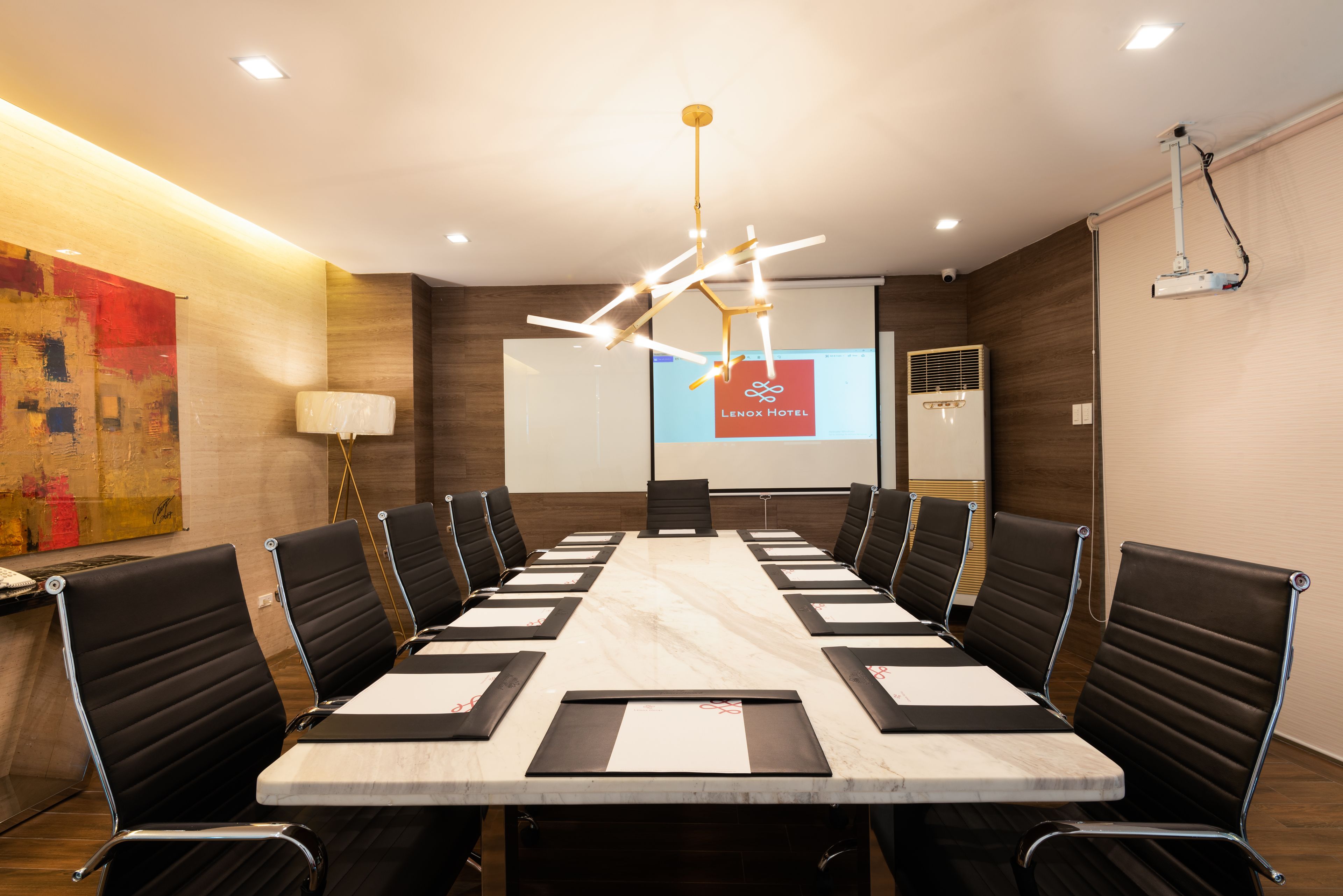 A conference room with a long table, black chairs, a projector screen, and a modern chandelier.