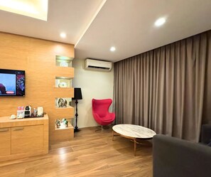 A modern hotel room with a wooden desk, a flat-screen TV, a red chair, a round table, and a large window with curtains.