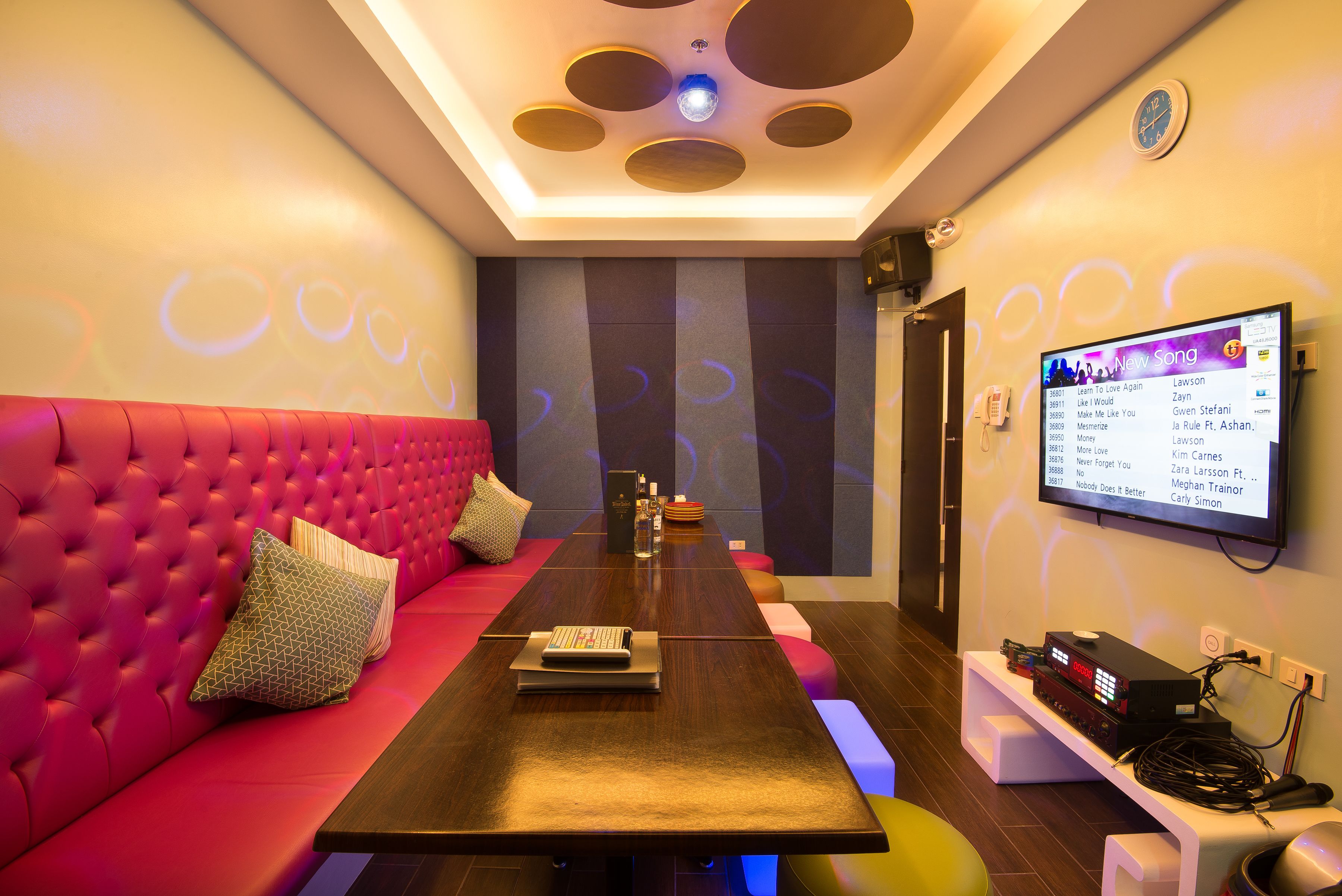 A modern lounge with a pink sofa, a long table, and a projection screen displaying a song list.