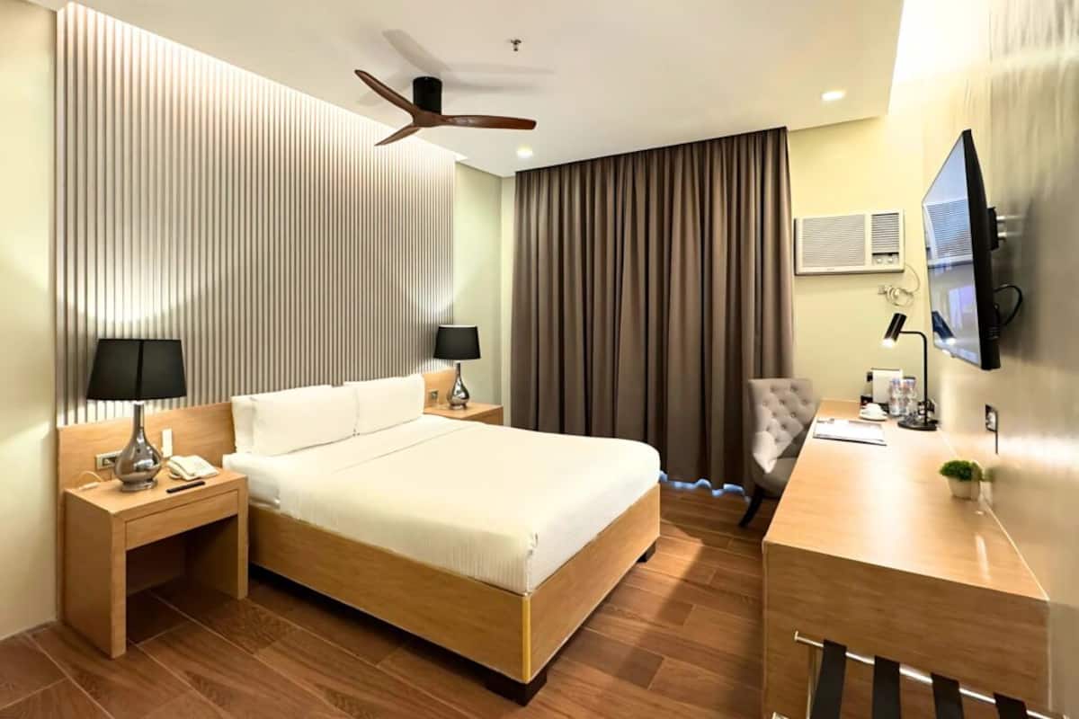 A modern hotel room with a large bed, a desk, a chair, a TV, and a ceiling fan.