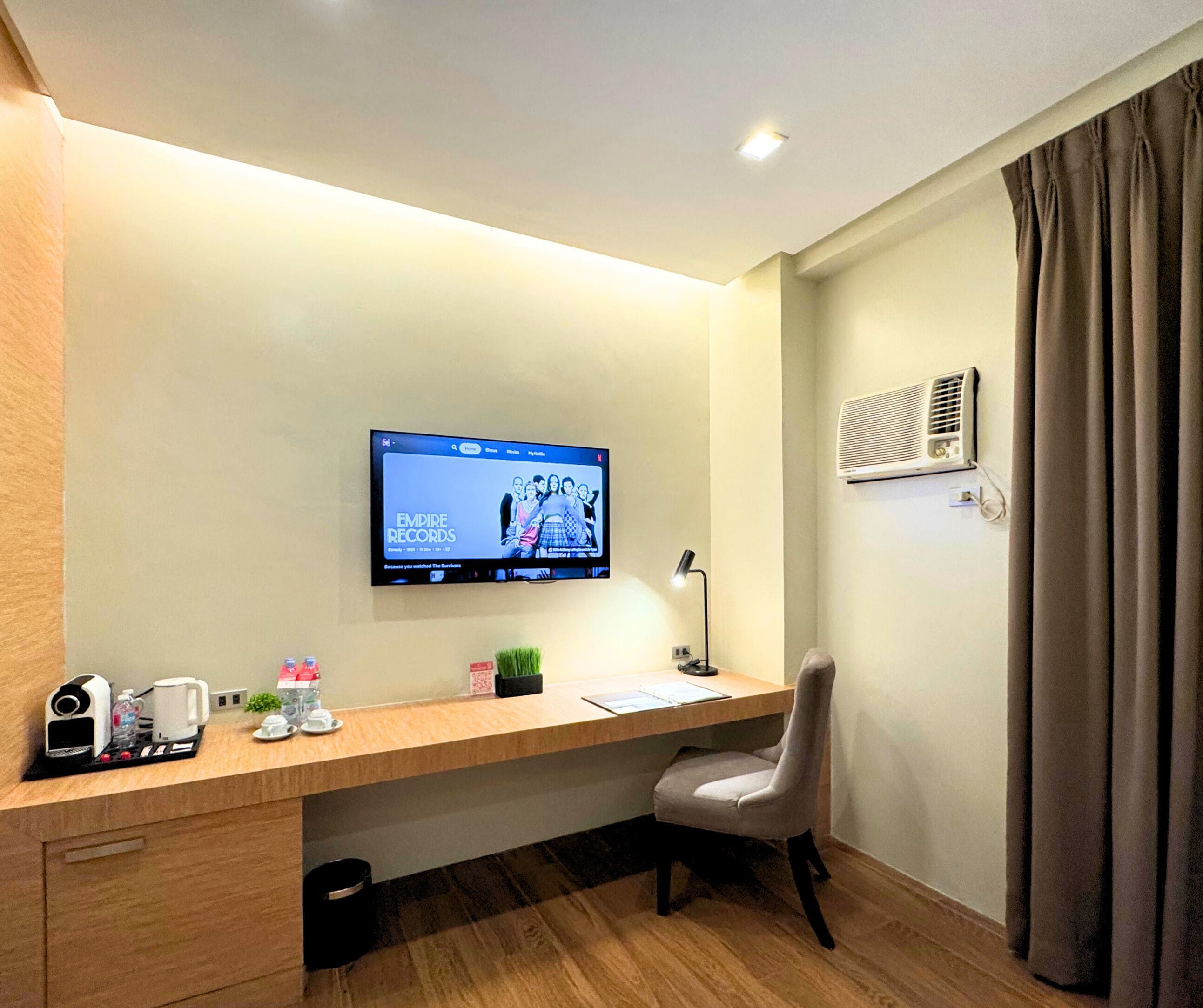 A hotel room with a wooden desk, a chair, a flat-screen TV mounted on the wall, a desk lamp, a coffee maker, a water bottle, a cup, a small plant, and an air conditioning unit.