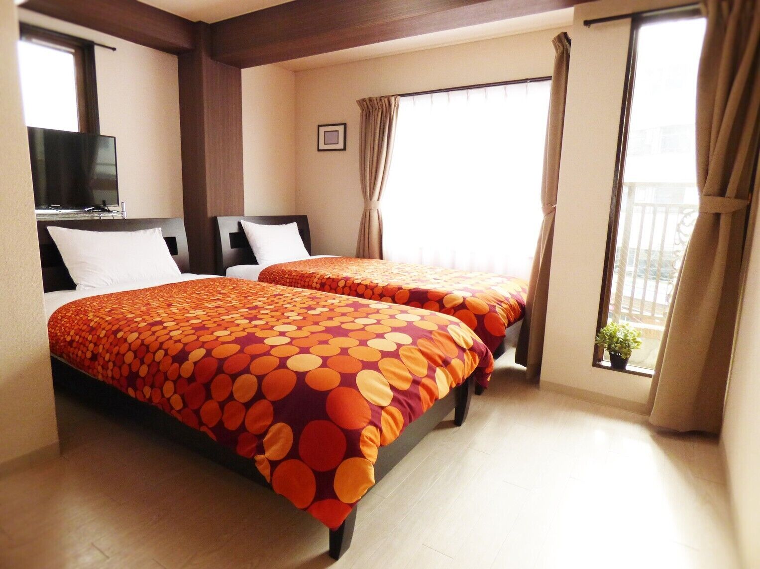 A hotel room with two beds, a TV, a window with curtains, and a balcony with a potted plant.